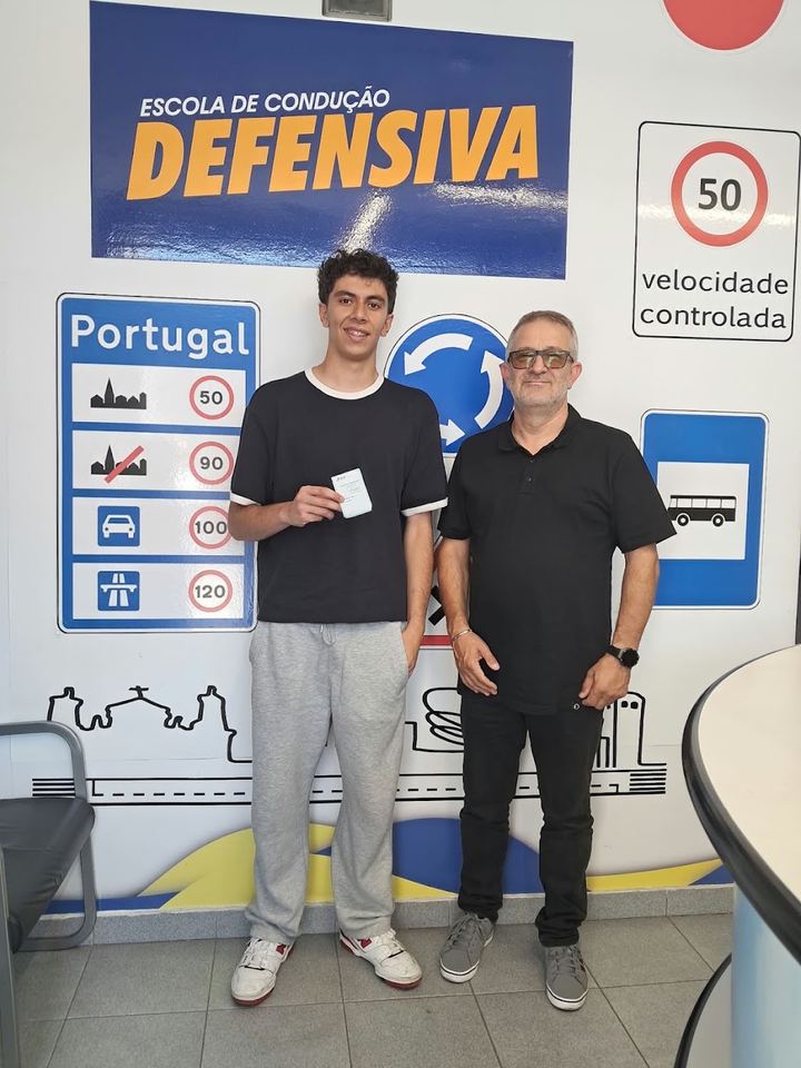 Novo condutor com o instrutor, a mostrar a carta em frente à parede de sinais da escola